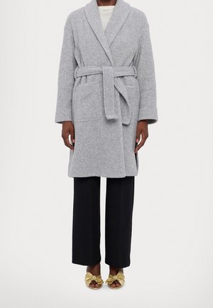 Personne portant un manteau gris ceinturé mi-long sur un pantalon noir et des talons ouverts dorés, debout devant un fond uni.