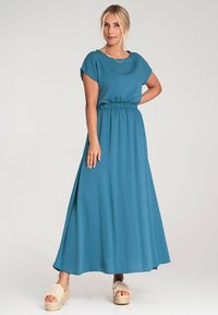 Turkusowa maxi sukienka z krótkimi rękawami, elastycznym paskiem w talii, zwiewnym materiałem o delikatnym połysku; zestawiona z sandałami na platformie posiadającymi teksturowany pasek.