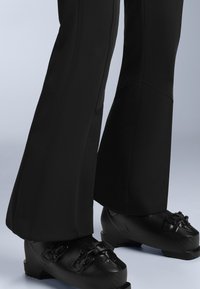 Fekete sí nadrág, harangláb stílusban, sima textúrával és kontrasztvarrással, erős fekete sícipőkkel, amelyeken állítható csatok vannak.