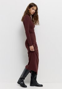 Abito fitted borgogna con maniche lunghe, realizzato in tessuto morbido; abbinato a stivali neri fino al ginocchio, che mettono in mostra una silhouette slanciata.
