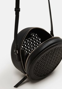 Borsa a tracolla rotonda nera in pelle con pannello anteriore intrecciato, zip parzialmente aperta e tracolla regolabile su uno sfondo semplice.