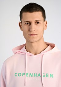 Ljusrosa hoodie i bomull, med en känguruficka och huva med dragsko. Tryckt text i grönt säger "COPENHAGEN INDBERGH."