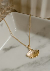 Collar de oro con un colgante en forma de hoja de ginkgo texturizada, que presenta una cadena lisa sobre un fondo de plato de mármol.