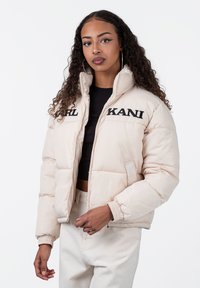 Beige dunjakke med høy krage, brodert "KARL KANI" tekst og elastiske mansjetter. Glatt, blank tekstur, glidelåslukking foran og sidelommer.