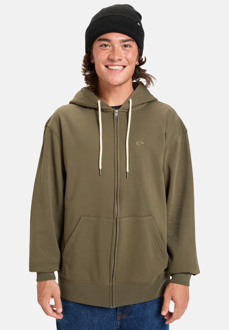 Sudadera con cremallera en verde oliva, con un bolsillo frontal, cordones en crema y un pequeño logo en el pecho. Tejido suave, corte relajado y mangas largas.