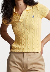 Gul, stickad polo med kabelmönster, krage och en liten blå logotyp på bröstet. Kortärmad och en åtsittande design.