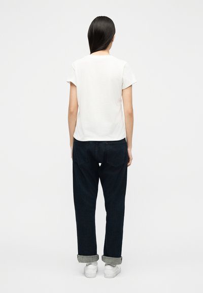 Γυναίκα με μακριά σκούρα μαλλιά, που στέκεται γυρισμένη με την πλάτη, φοράει λευκό κοντομάνικο πουκάμισο, σκουρόχρωμο τζιν με ψηλόμεσο καμπάνα που είναι τυλιγμένο και λευκά αθλητικά παπούτσια, σε απλό φόντο.