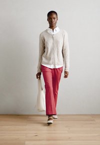 Cardigan beige clair sur une chemise blanche, associé à un pantalon rose et des chaussures crème. Tenant un sac blanc, debout sur un sol en bois.