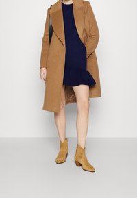 Person klädd i en marinblå klänning med volangkant, lång kappa i kamelfärg, tan mocka ankelskor och en svart axelremsväska mot en vit bakgrund.