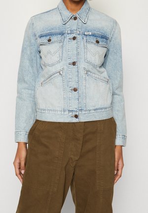 Person iført lyseblå denimjakke med knapper og bryst- og sidelommer, kombineret med brune bukser med høj talje.