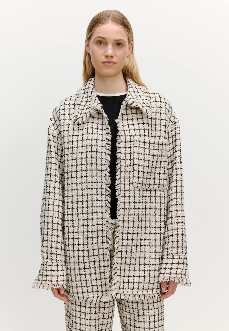 Tweed jasje in een crèmekleurig en zwart geruit patroon, met een franjeszoom, oversized pasvorm en grote kraag. G gedragen over een zwart top.