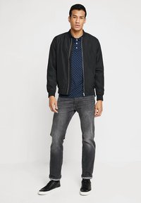Černá bomber bunda se zipem vpředu, spárovaná s námořnickou polo košile s puntíky a šedými džíny. Černé tenisky doplňují celý outfit.