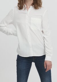 Chemise blanche boutonnée avec un col pointu, des manches longues et une poche poitrine, confectionnée dans un tissu lisse avec une texture légèrement brillante.