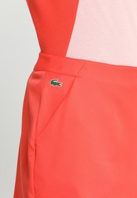 Koralejowa sukienka z dopasowanym krojem, gładką teksturą i detalem kieszeni. Zawiera mały akcent w postaci zielonego logo krokodyla.