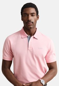 Muž s krátkými kudrnatými vlasy a knírem, který má na sobě světle růžové polo tričko a stříbrné hodinky, stojí před jednobarevným bílým pozadím.