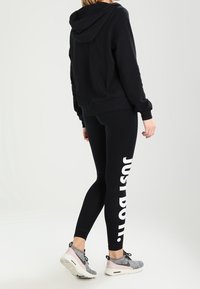 Person wearing black hoodie and black leggings with white "JUST DO IT." text, paired with gray and pink athletic shoes, facing away.