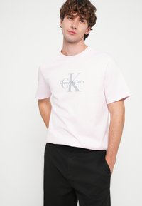 Ανοιχτό ροζ βαμβακερό μπλουζάκι με κεντρικό γκρι λογότυπο με την επιγραφή «Calvin Klein». Κοντά μανίκια και στρογγυλή λαιμόκοψη. Συνδυάζεται με μαύρο παντελόνι.