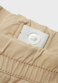 Beige tyg med fin ribbning, med en vit textilflik som har en rund knapp och tät sömnad längs kanterna.