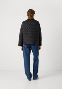 Jachetă neagră matlasată cu guler din catifea maro. Spatele are o croială dreaptă și este asortată cu blugi albaștri.