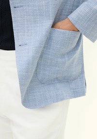 Blazer a righe azzurre presenta una vestibilità rilassata, con una grande tasca frontale e un tessuto testurizzato su un outfit bianco.