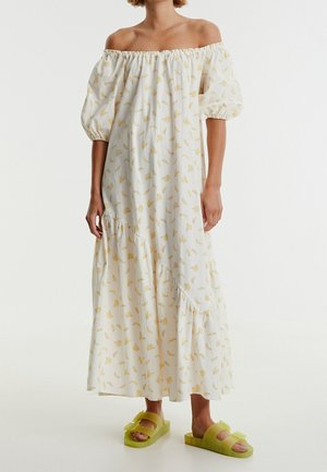 Femme portant une robe maxi blanche épaules dénudées avec imprimé floral jaune et manches bouffantes, combinée à des sandales slides jaunes avec nœuds.