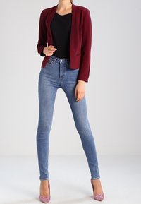 Blazer bordeaux porté sur un haut noir, associé à un jean skinny bleu clair et des escarpins pailletés roses. Textures lisses avec un minimum de détails métalliques.
