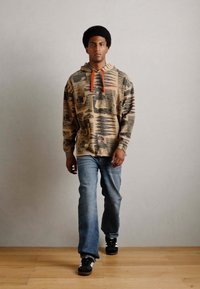 Hooded sweatshirt with tan base and black graphic prints. Orange drawstrings, paired with light blue jeans and black sneakers.