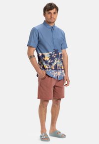 Blå kortärmad skjorta med knappar och palmträdsgrafik i gult, tillsammans med rostfärgade shorts och ljusblå sandaler.