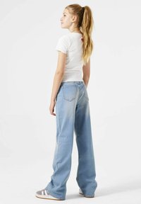 Fittend wit geribbelde t-shirt met rood logo, gecombineerd met lichtblauwe wijde jeans. De jeans hebben twee achterzakken en subtiele vervaging.