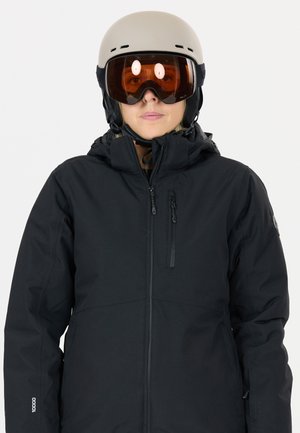 Veste isolante noire avec un col haut, une fermeture éclair sur le devant et plusieurs poches. Associée à un casque beige et de grandes lunettes de ski marron.
