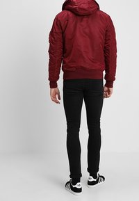 Bordeauxrode donsjas met capuchon, geribbelde manchetten en zoom, gestructureerde mouwen. Draagt over zwarte slim-fit jeans en zwart-wit gestreepte sneakers.