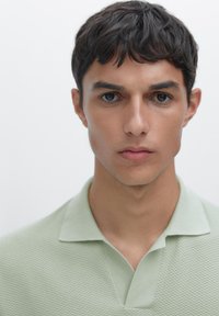 Polo shirt verde chiaro con texture, colletto classico e un delicato design a costine, indossato da una persona con capelli scuri e corti e un'espressione neutra.