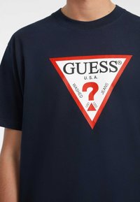 T-shirt in cotone blu navy con un grande logo triangolare rosso e bianco con la scritta "GUESS," un punto interrogativo e il testo "WASHED JEANS."