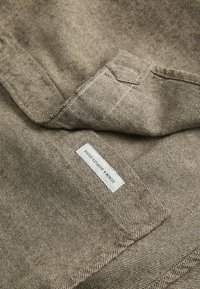 Bruine gestreepte stof met een zachte textuur, voorzien van een label met "Denim & Supply Goods" in het wit. De stof is gevouwen, waarbij het patroon zichtbaar is.