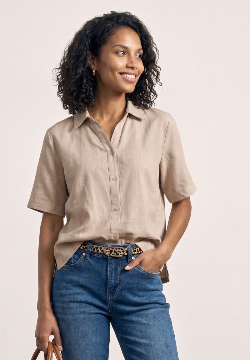 Vrouw met krullend haar die een beige kort mouw blouse met knopen draagt, blauwe jeans, een luipaardprint riem, een bruine leren tas vasthoudt, glimlacht en naar rechts kijkt.