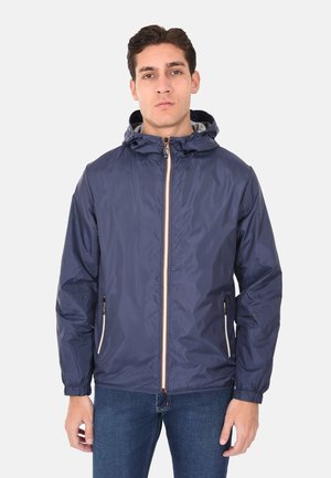 Giovane uomo che indossa una giacca a vento blu navy con cappuccio e zip, con tasche con cerniera e polsini elastici, abbinata a jeans blu.