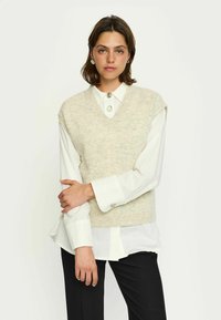 Beige kötött mellény V-nyakkal, fehér galléros ing fölött viselve. Rövid ujjakkal rendelkezik és puha tapintású, fekete nadrággal párosítva.