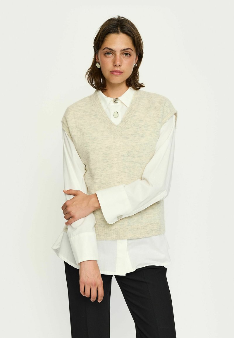 Beige kötött mellény V-nyakkal, fehér galléros ing fölött viselve. Rövid ujjakkal rendelkezik és puha tapintású, fekete nadrággal párosítva.