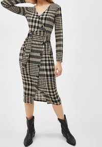 Svart och beige rutigt omslagsklänning med långa ärmar, V-ringning, knappdetaljer, knytmidja och knälång nederdel. Kombineras med svarta stövlar.