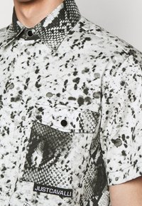 Camicetta a maniche corte in bianco e nero, con una stampa a serpente testurizzata, motivi puntinati e una tasca frontale con un dettaglio del logo.
