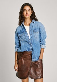 Denim srajca z gumbi in žepi na prsih, kombinirana z rjavim usnjenim ovojem, ki ima gladko teksturo in stranske žepe.