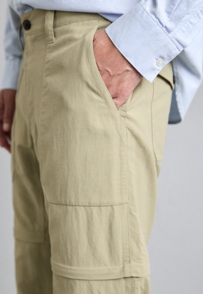 Un pantalon cargo beige en tissu lisse et texturé. Doté de poches latérales et d'une coupe décontractée. Associé à une chemise à manches longues bleu clair.