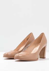 Escarpins en cuir verni beige avec un bout arrondi et un talon large, mettant en avant une texture lisse et des détails minimalistes.