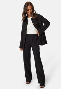 Zwarte jas met knopen en een ontspannen pasvorm over een witte top, gecombineerd met zwarte, ruime broek. Accessoires zijn zwarte sandalen.