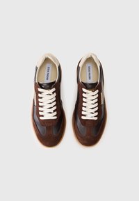Sneakers din suede și piele maro, cu șireturi albe, vârf rotund și talpă din cauciuc bej. Prezintă eticheta brandului pe branț.