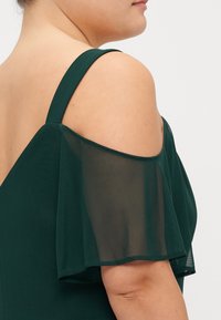 Abito verde scuro con maniche a sbuffo trasparenti e scollatura fredda, texture liscia e design aderente del corpetto. Dettagli di cucitura sottili ai bordi.