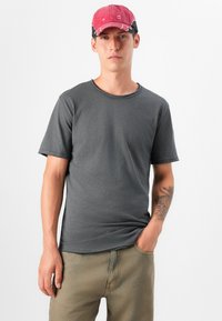 Un joven con un tatuaje lleva una camiseta gris y una gorra rosa, de pie frente a un fondo blanco, con las manos en los bolsillos.