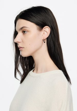 Mudel, kellel on pikad tumedad juuksed ja seljas kreemivärvi pullover. Müravad kuldsed rõngasseppe kontrastiks riide pehme tekstuuriga.