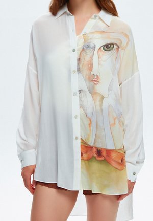 Femme portant une chemise blanche transparente boutonnée avec des motifs abstraits de visages et des fleurs orange sur le devant, associée à un short marron.