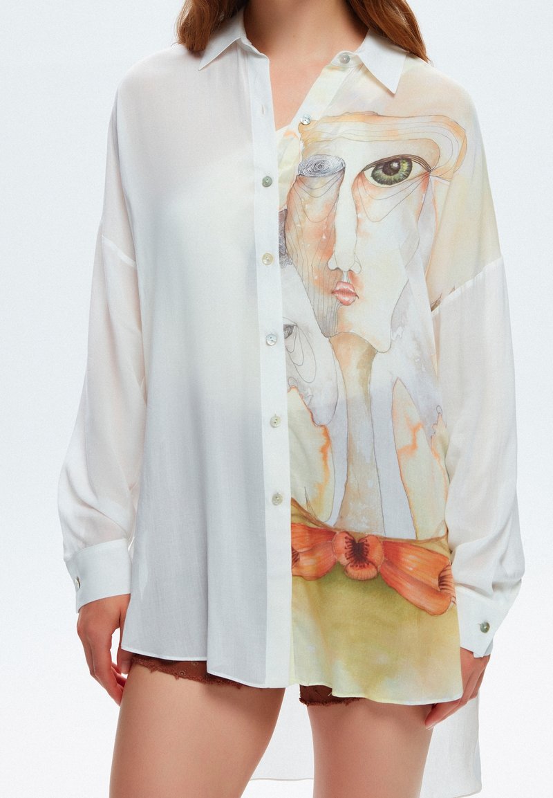 Femme portant une chemise blanche transparente boutonnée avec des motifs abstraits de visages et des fleurs orange sur le devant, associée à un short marron.